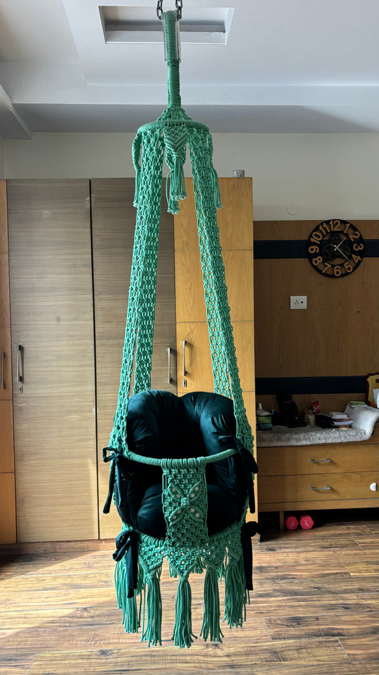 Green macrame hammock with black cushion hanging in a room.