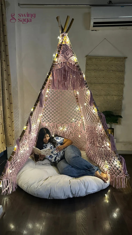 macrame teepee in pink color decorated with fairy lights with cushion and mattress in which a girl is reading a book placed in a room setting with wooden flooring.