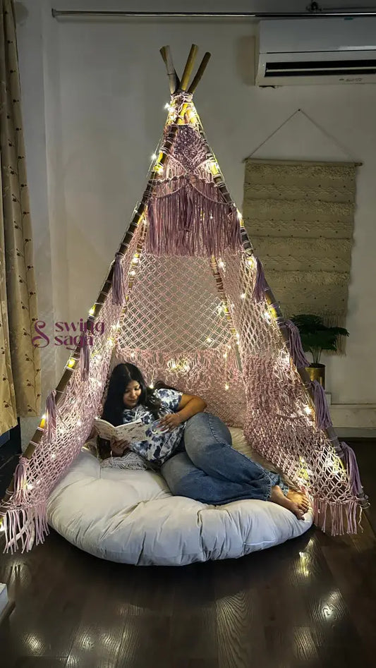 macrame teepee in pink color decorated with fairy lights with cushion and mattress in which a girl is reading a book placed in a room setting with wooden flooring.