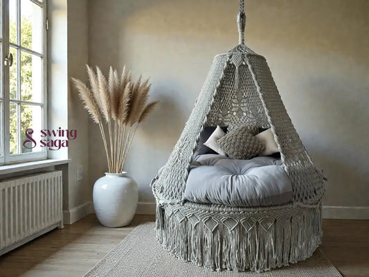 Grey colored macrame swing with mattress for sitting in an indoor room corner with rug.