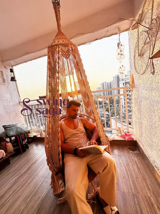 Man reading a book on a handmade beige macrame swing chair in a high-rise balcony during sunset, with a city skyline in the background.