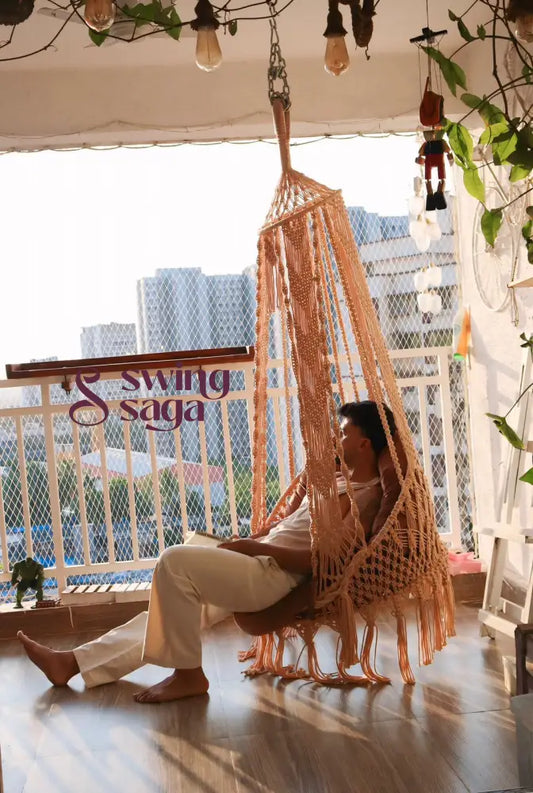 Man sitting on a handcrafted beige macrame timber chair swing, swing on a high-rise balcony at sunset, with city buildings in the background.