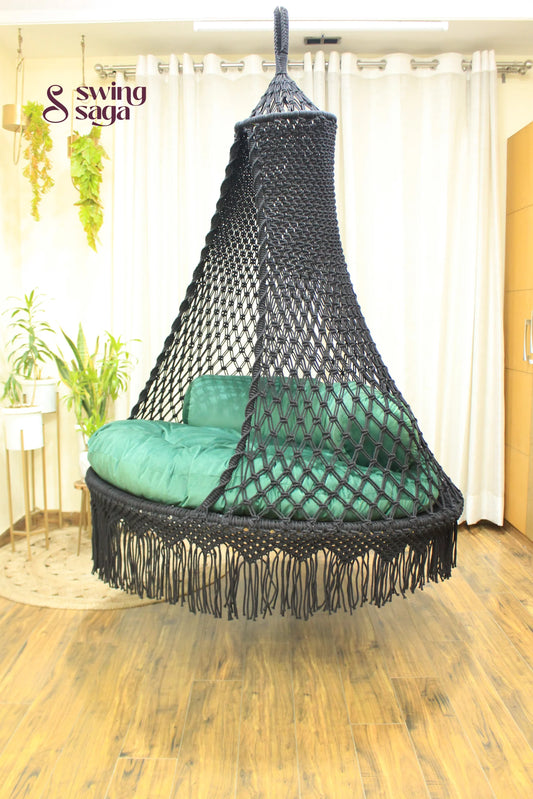 Black hanging macrame swing in a room with white cutains, planters and wooden flooring.