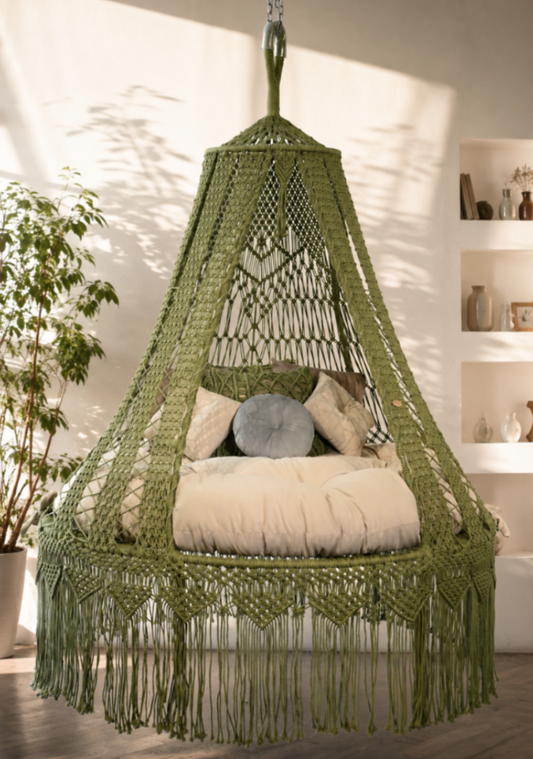 An olive green macrame hanging swing chair with fringe, styled with cream cushions and pillows in a bright, modern room with warm sunlight and shadows.