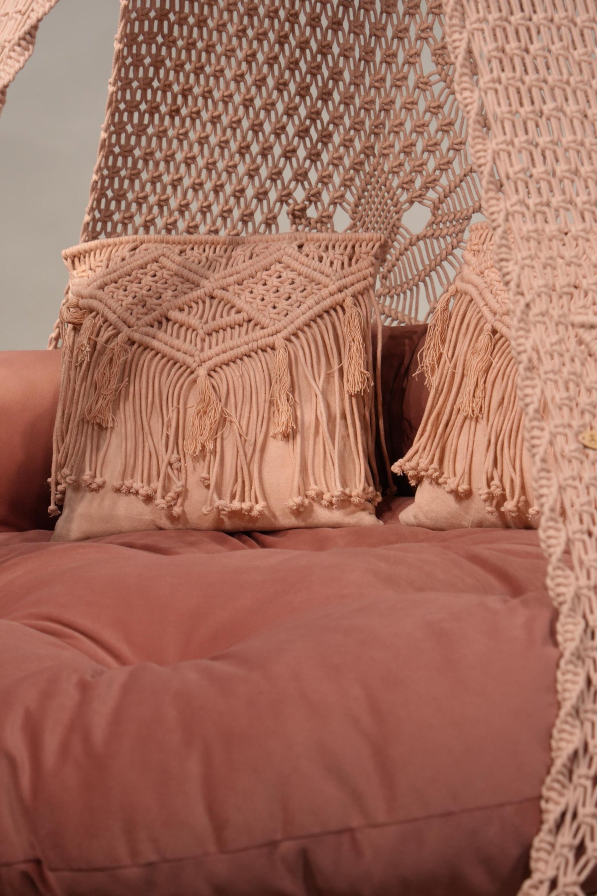 A close-up of a dusty rose velvet sofa cushion styled with matching beige macrame pillows and a woven textile hanging in the background.