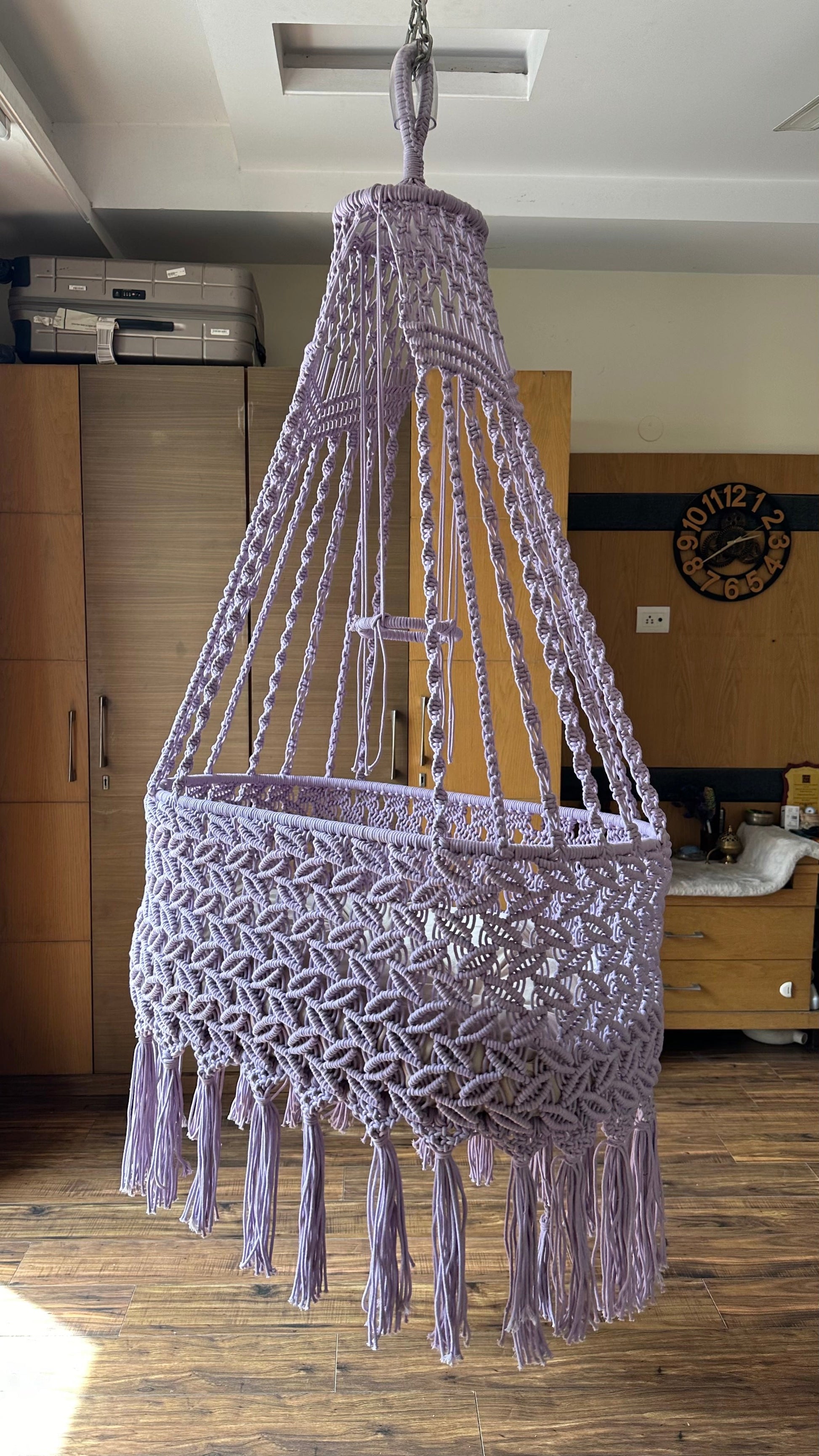 Handmade lavender macrame baby bassinet hanging in a nursery room with soft cushion.
