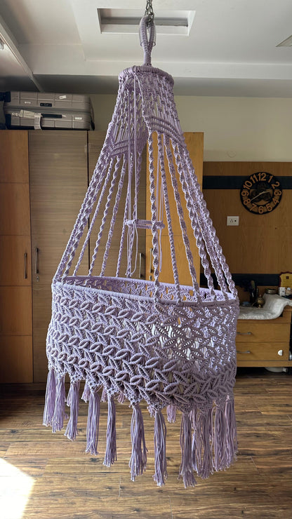 Handmade lavender macrame baby bassinet hanging in a nursery room with soft cushion.