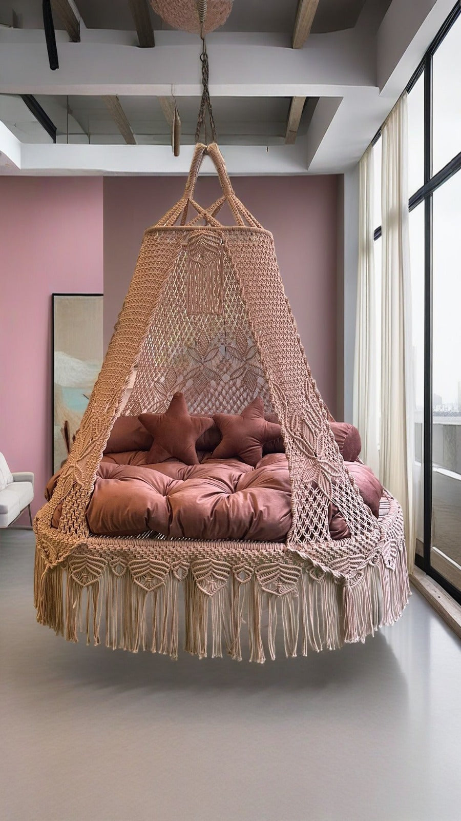Hanging macrame bed with pink cushions in a room with pink walls and large windows.