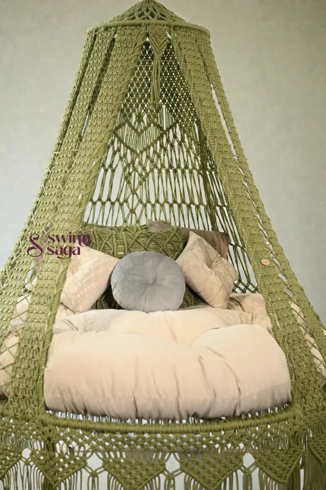 Green macramé hanging swing with fringe, beige cushion, and pillows placed in a bright room with large windows and balcony view.