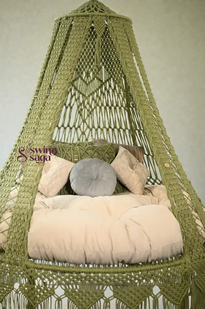 Green macramé hanging swing with fringe, beige cushion, and pillows placed in a bright room with large windows and balcony view.