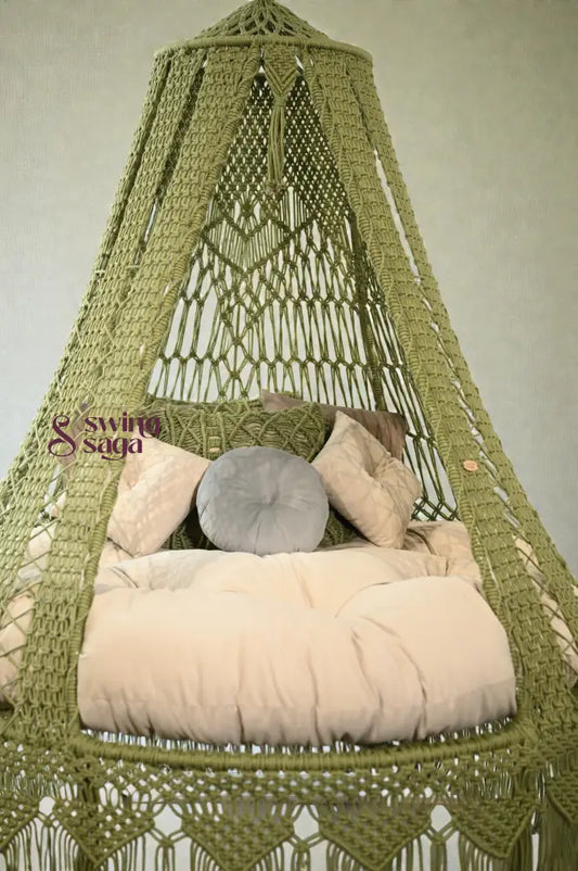Green macramé hanging swing with fringe, beige cushion, and pillows placed in a bright room with large windows and balcony view.