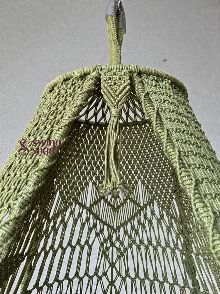 Close-up view of an olive green handcrafted macrame swing top, showing detailed knotting, braided hanging loop, and netted pattern against a neutral wall background.