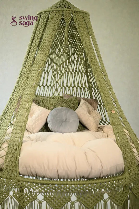 Green macramé hanging swing with fringe, beige cushion, and pillows placed in a bright room with large windows and balcony view.