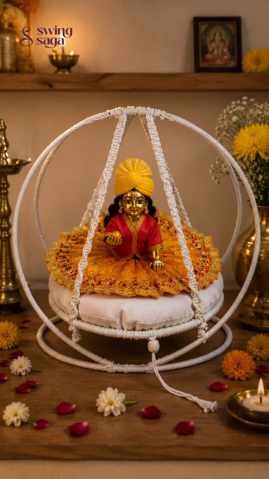 A decorative white circular hanging cradle or swing cradle holding a small gold-toned deity statue dressed in red and yellow, placed on a wooden surface with flowers, candles, and brass lamps around it.