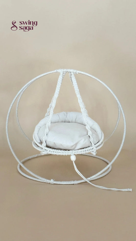 A white macrame hanging chair with a round woven frame, suspended seat cushion, and matching floor loop, photographed against a plain beige studio background.