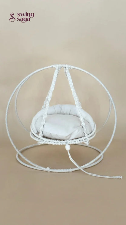 A white macrame hanging chair with a round woven frame, suspended seat cushion, and matching floor loop, photographed against a plain beige studio background.