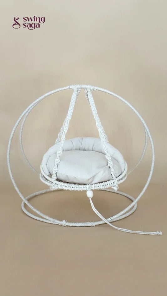 A white macrame hanging chair with a round woven frame, suspended seat cushion, and matching floor loop, photographed against a plain beige studio background.