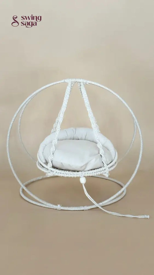 A white macrame hanging chair with a round woven frame, suspended seat cushion, and matching floor loop, photographed against a plain beige studio background.