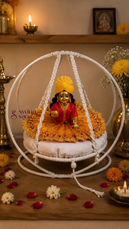 A decorative white circular hanging cradle or swing cradle holding a small gold-toned deity statue dressed in red and yellow, placed on a wooden surface with flowers, candles, and brass lamps around it.