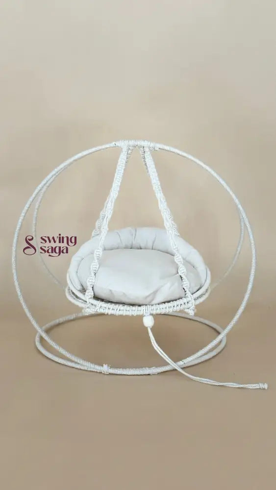 A white macrame hanging chair with a round woven frame, suspended seat cushion, and matching floor loop, photographed against a plain beige studio background.