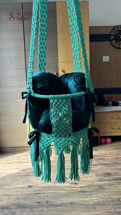 A dusky teal baby swing made of cotton, with a decorative tassel design, hanging indoors.