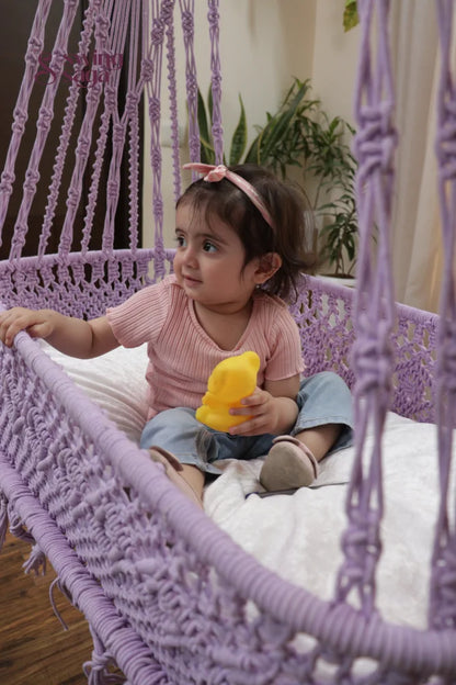 Cute baby girl sitting in a handwoven lavender macrame cradle swing holding a to