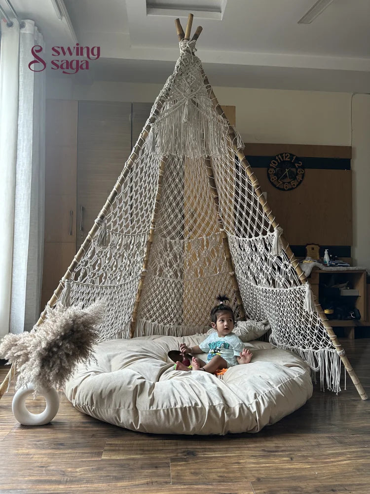 A baby is sitting in a white macrame teepee placed on a wooden floor.