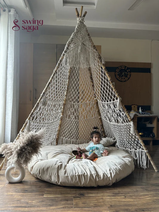 A baby is sitting in a white macrame teepee placed on a wooden floor.