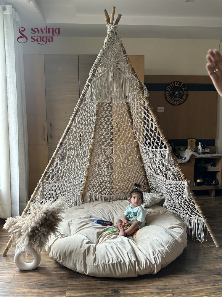 A baby is sitting in a white macrame teepee placed on a wooden floor.
