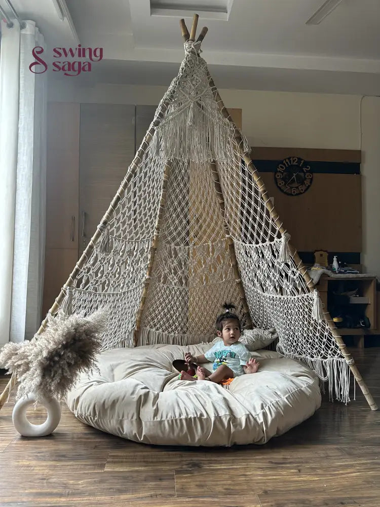 A baby is sitting in a white macrame teepee placed on a wooden floor.