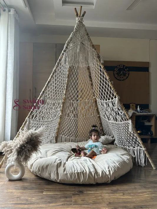 A baby is sitting in a white macrame teepee placed on a wooden floor.