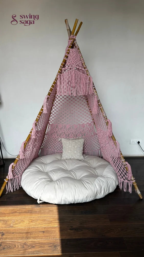 Pink macrame teepee with white mattress and a tiny square cushion placed on a wooden floor.