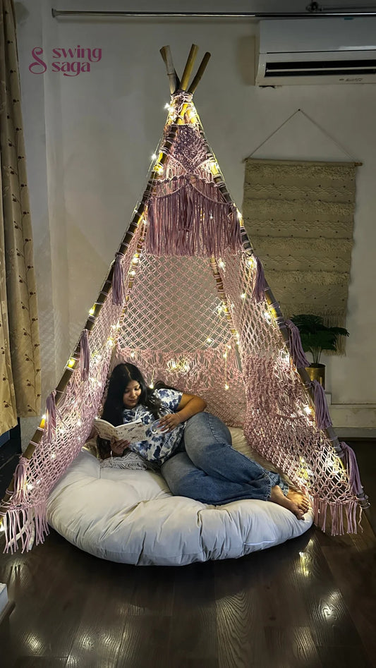 macrame teepee in pink color  decorated with fairy lights with cushion and mattress in which a girl is reading a book placed in a room setting with wooden flooring.