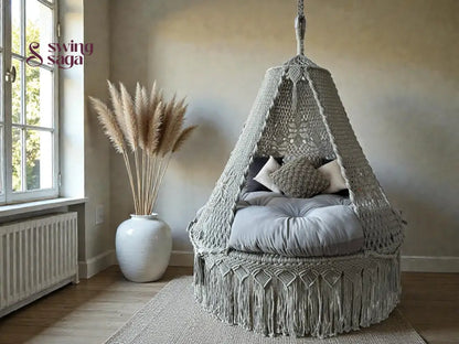 Grey colored macrame swing with mattress for sitting in an indoor room corner with rug. 