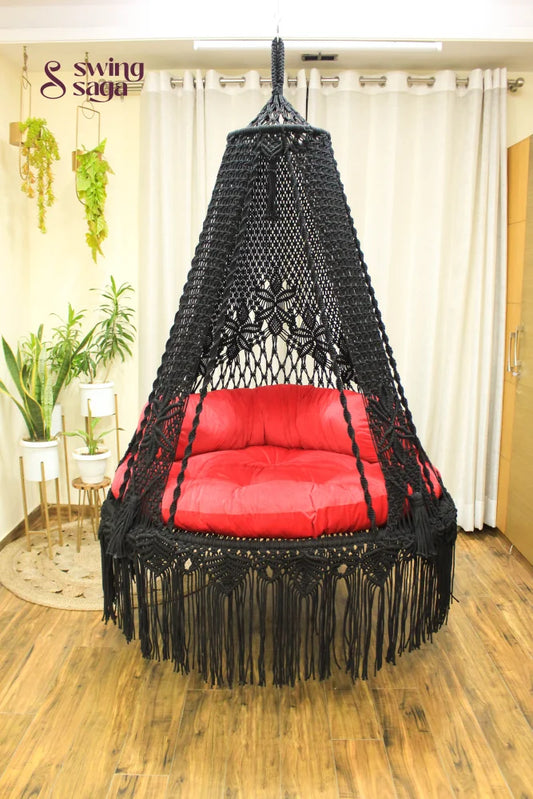 Handmade macrame swing in black color, hanging in a room with white curtains and wooden flooring.