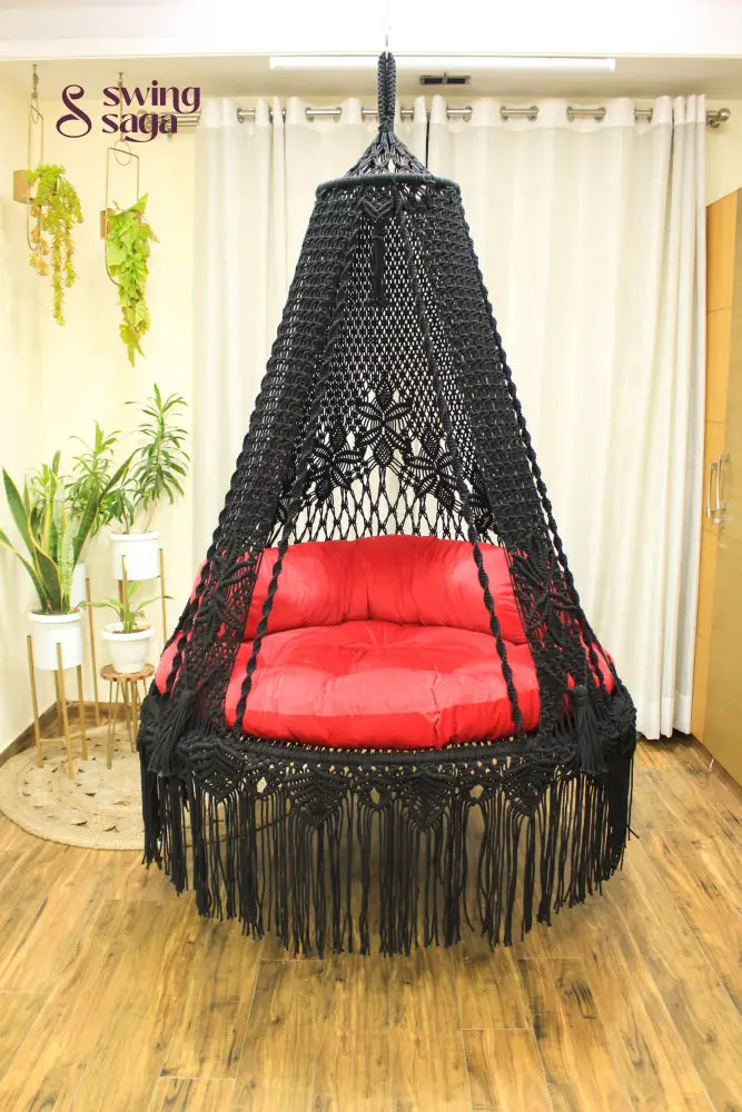 Handmade macrame swing in black color, hanging in a room with white curtains and wooden flooring.