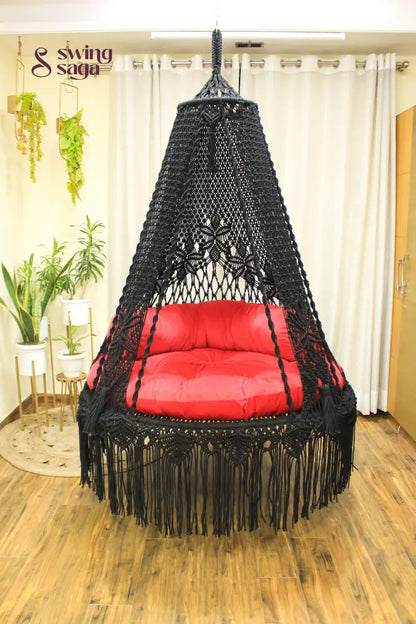 Handmade macrame swing in black color, hanging in a room with white curtains and wooden flooring.