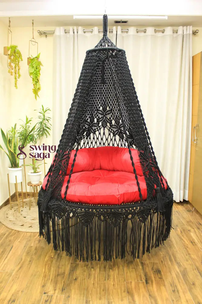 Handmade macrame swing in black color, hanging in a room with white curtains and wooden flooring.