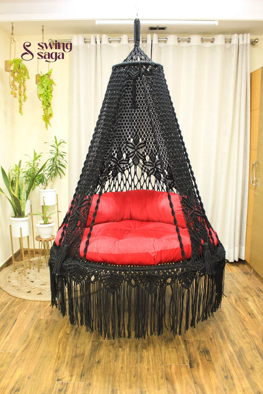 Handmade macrame swing in black color, hanging in a room with white curtains and wooden flooring.