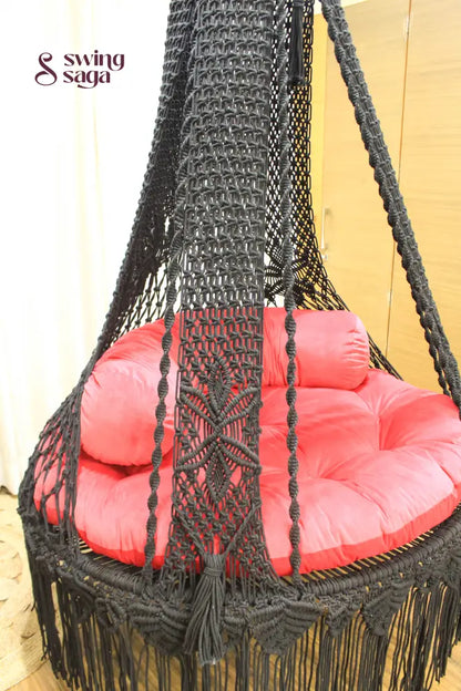 Macrame hanging swing with red mattress and cylindrical cushion.