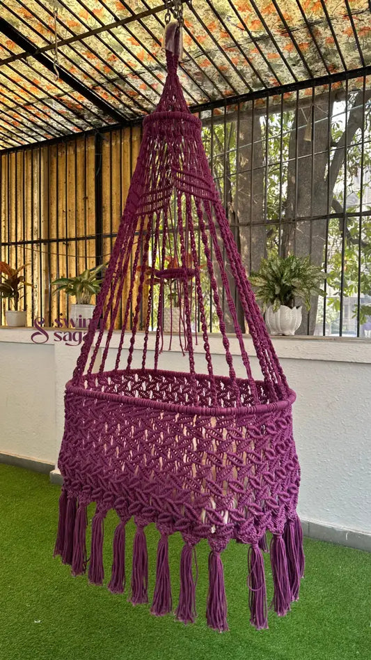 Purple macrame hanging chair swing with tassel fringe, suspended indoors in a modern glass-window home setting.