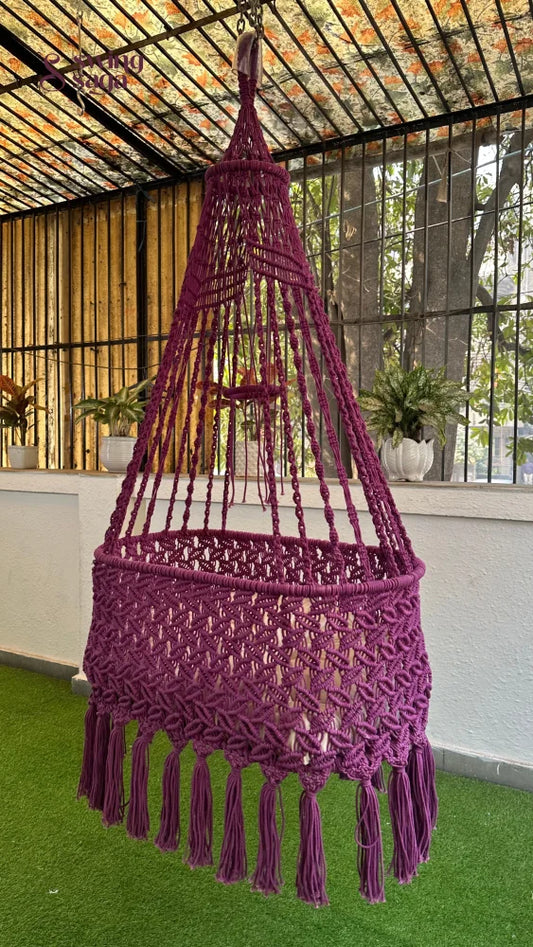 Purple macrame hanging chair swing with tassel fringe, suspended indoors in a modern glass-window home setting.