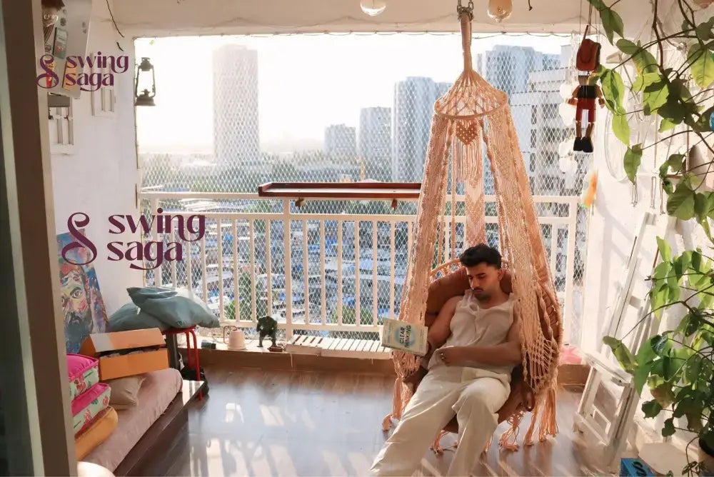 A guy in a balcony area sitting on a macrame swing reading a book.