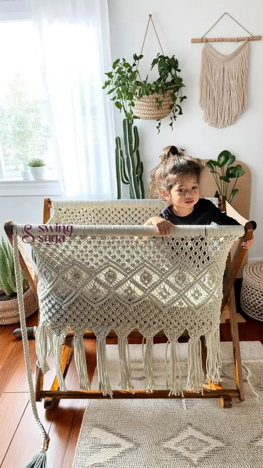 Child playing in a white macrame bassinet in a bright room with plants.