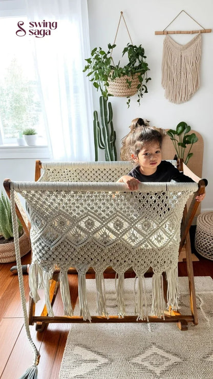 Child playing in a white macrame bassinet in a bright room with plants.