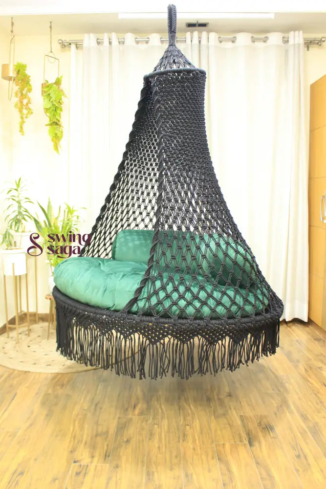 Black hanging macrame swing in a room with white cutains, planters and wooden flooring.