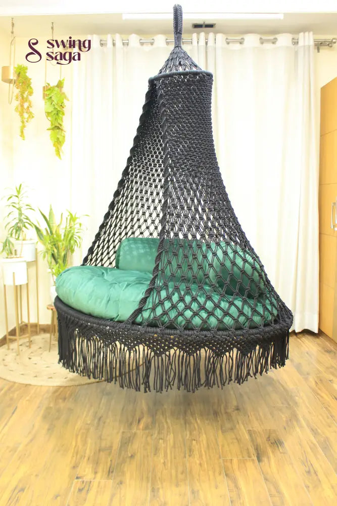 Black hanging macrame swing in a room with white cutains, planters and wooden flooring.